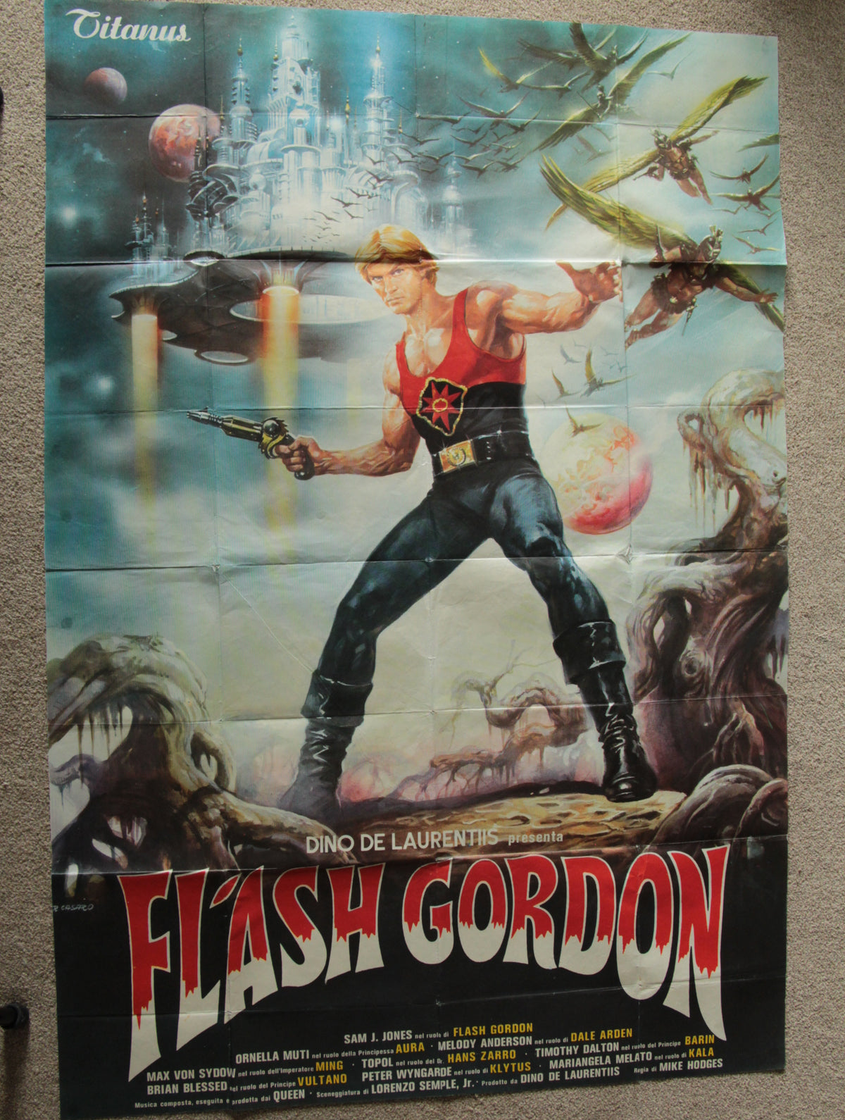 A vintage Flash Gordon movie poster displayed flat against a neutral background. The poster features the character Flash Gordon in a red and black costume, surrounded by sci-fi creatures, spaceships, planets, and a castle in the background, with the title 'FLASH GORDON' in large red letters at the bottom.